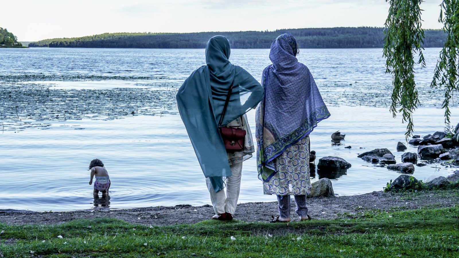 Bild på tå kvinnor framför en sjö där ett barn doppar fötterna