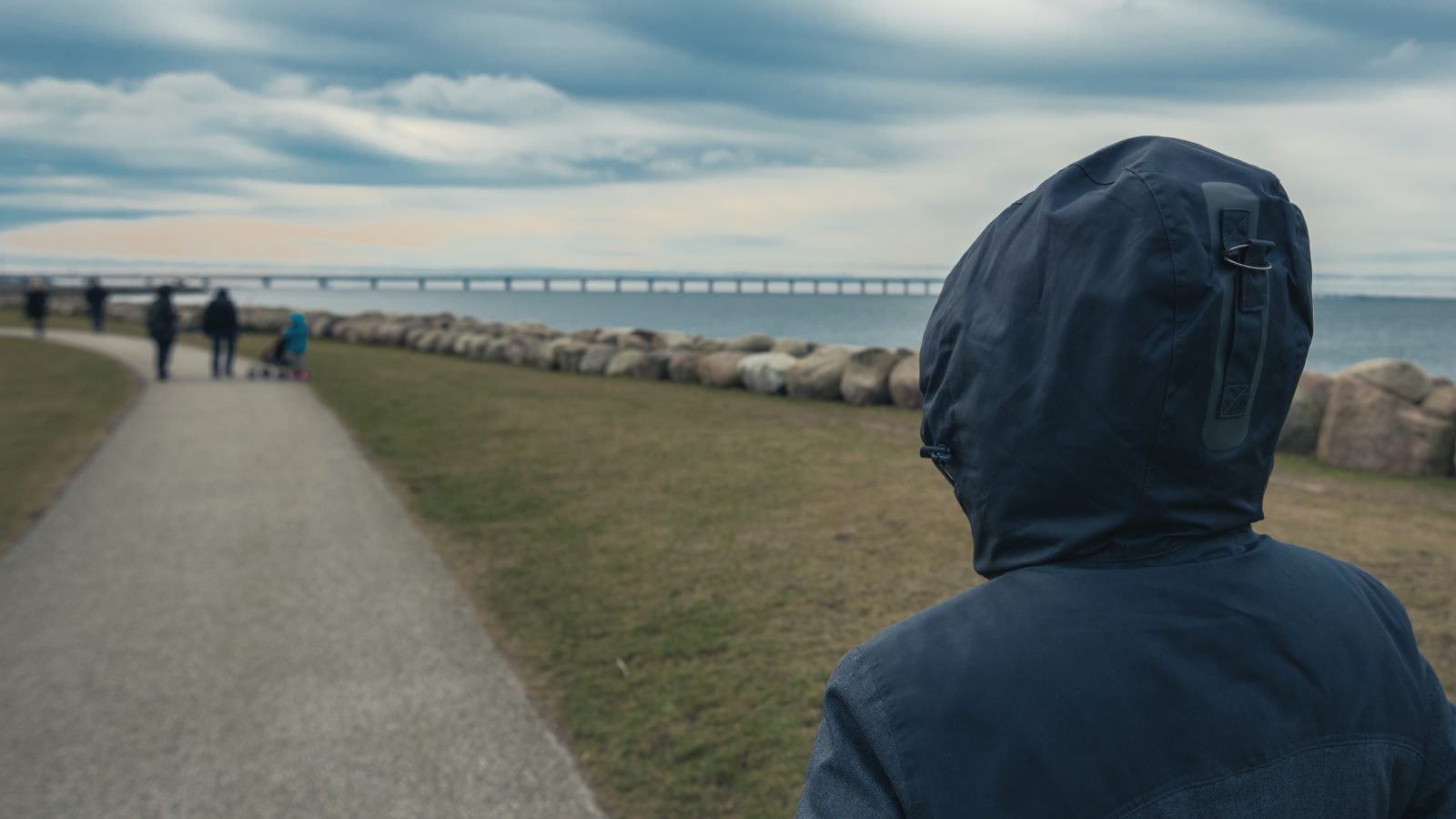 Person med hoodie står vid vattnet med en bro på avstånd