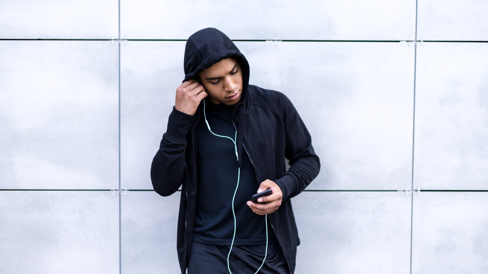 Kille med hoodie står lutad mot vägg och sätter in sina hörlurar i örat