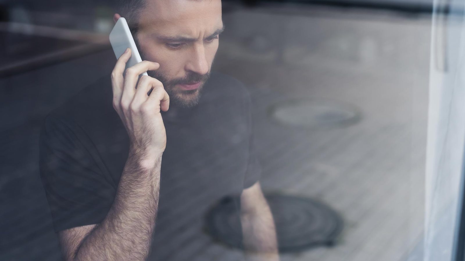Allvarlig man ståendes vid fönster med mobil i handen