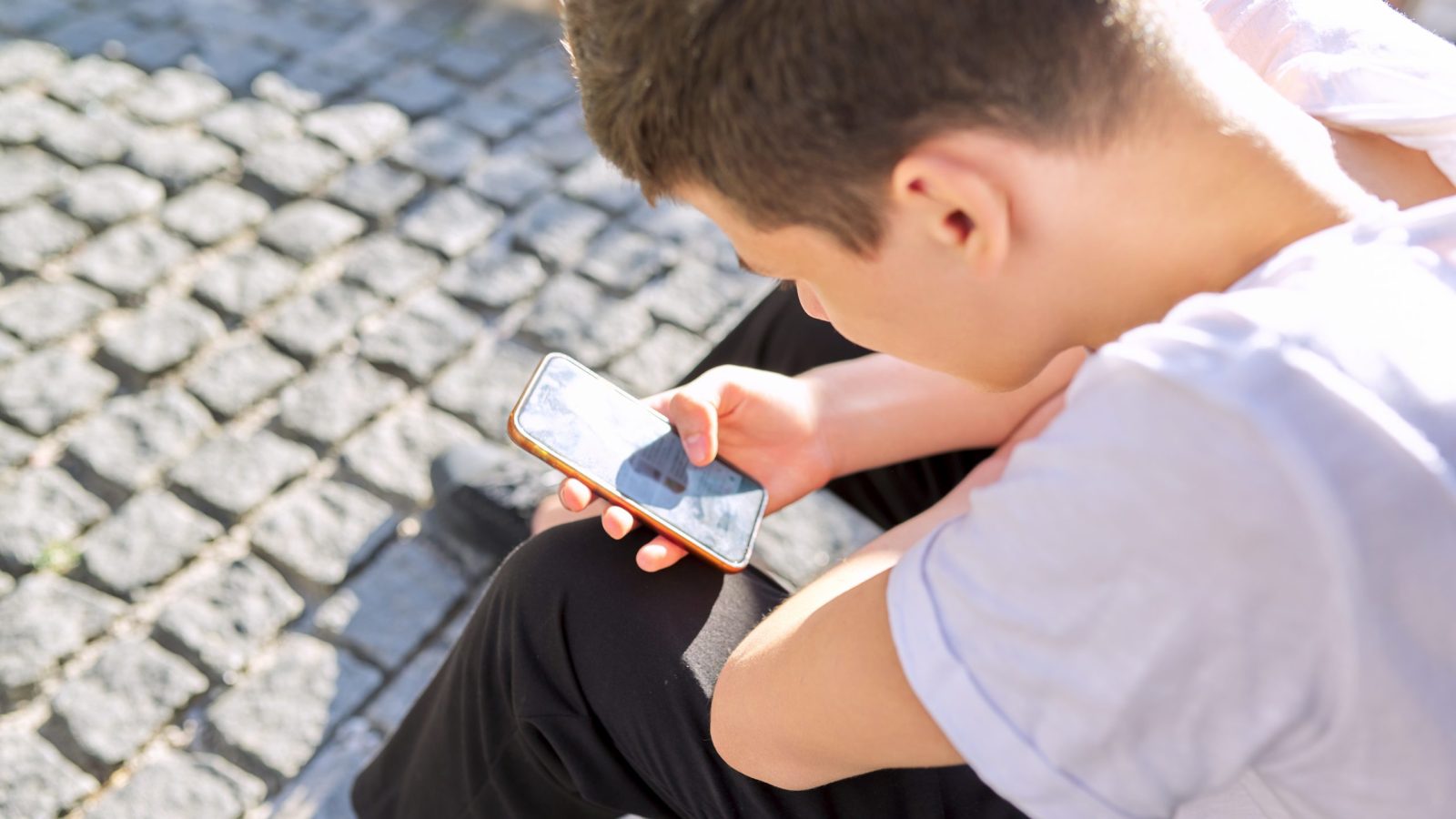 ung kille som sitter på en kullerstensgata ed mobiltelefonen i höger hand