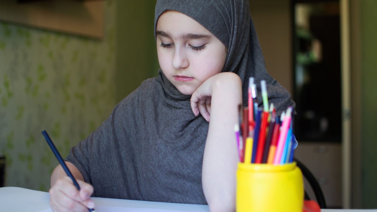 flicka i 11-12 års åldern ikläddi grå hijab som sitter vid ett skrivbord och ritar