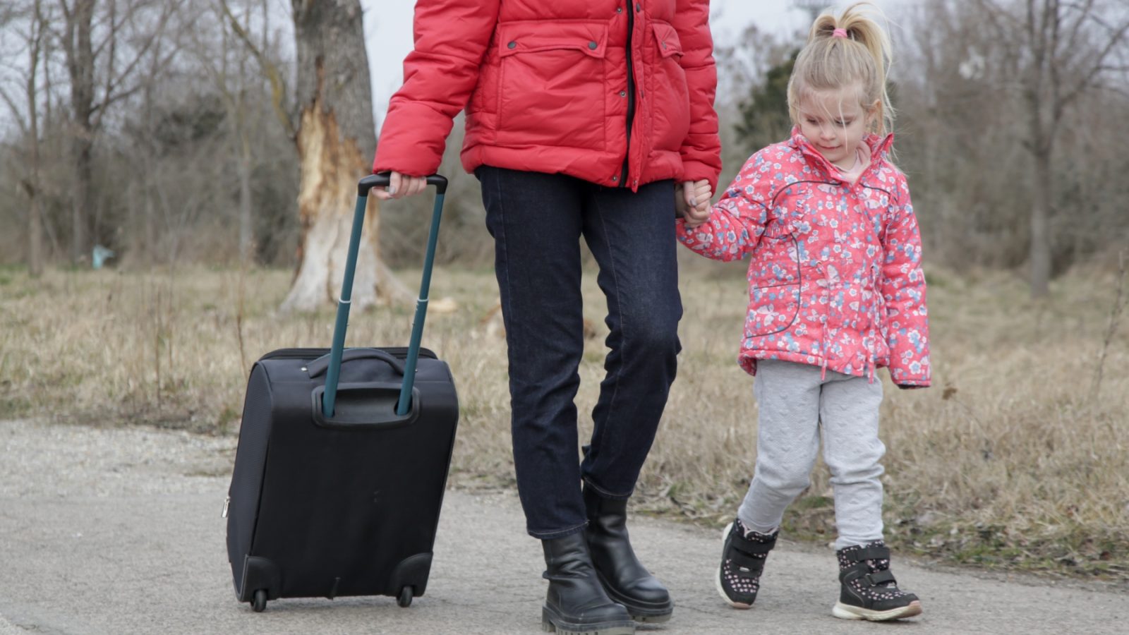 Woman holding suitcase in one hand and child in the other in seen walking.