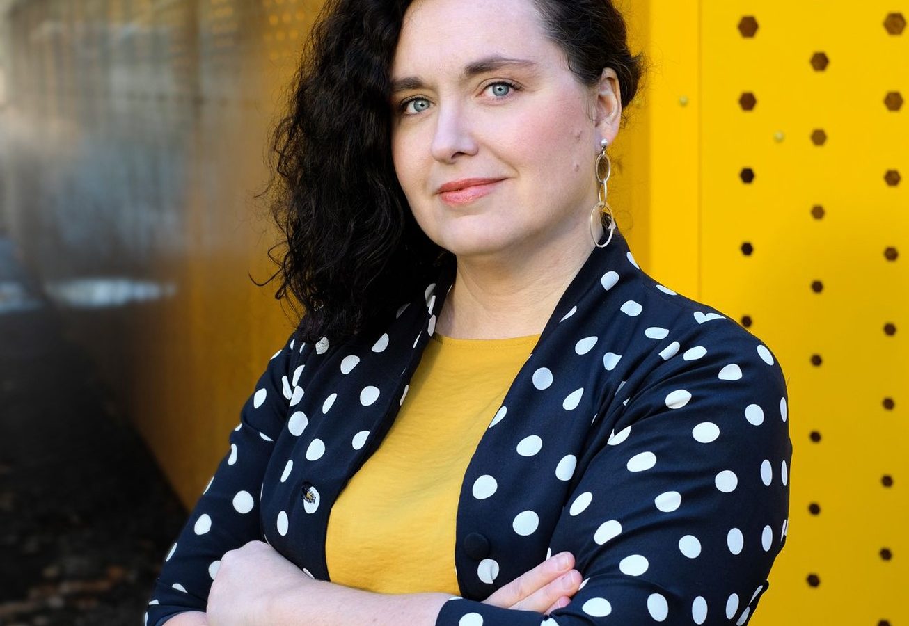 Foto av Olga Szczepankiewicz som har mörkt hår, prickig blus med gul tröja under och står med armarna i kors.