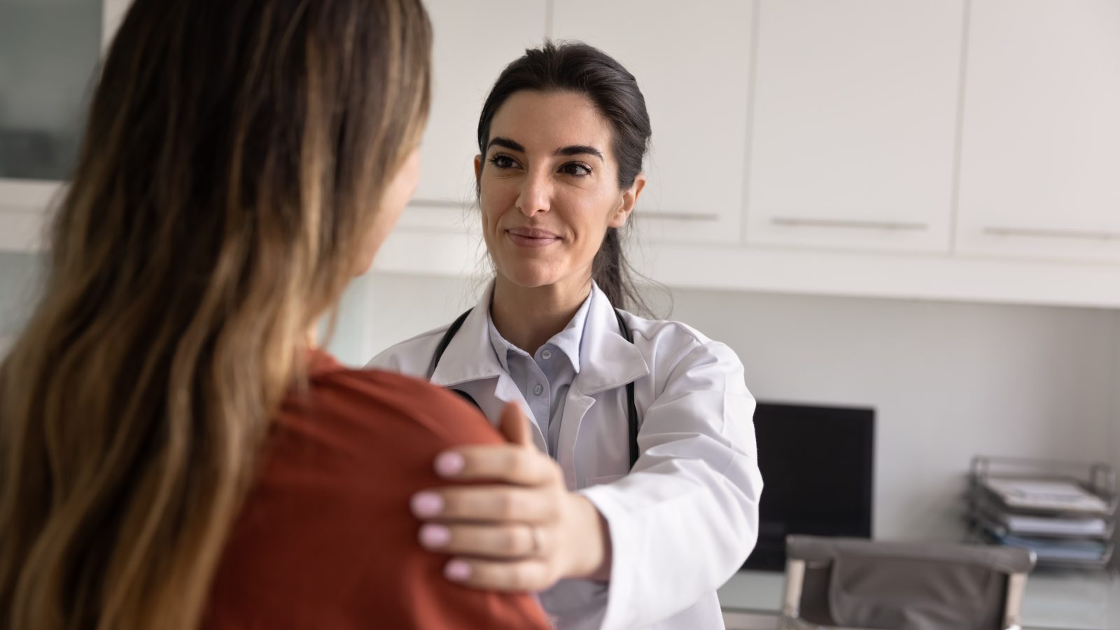 Positive satisfied young Latin doctor woman touching shoulder of patient with support, discussing good healthcare news, medical checkup result, successful therapy ending, smiling